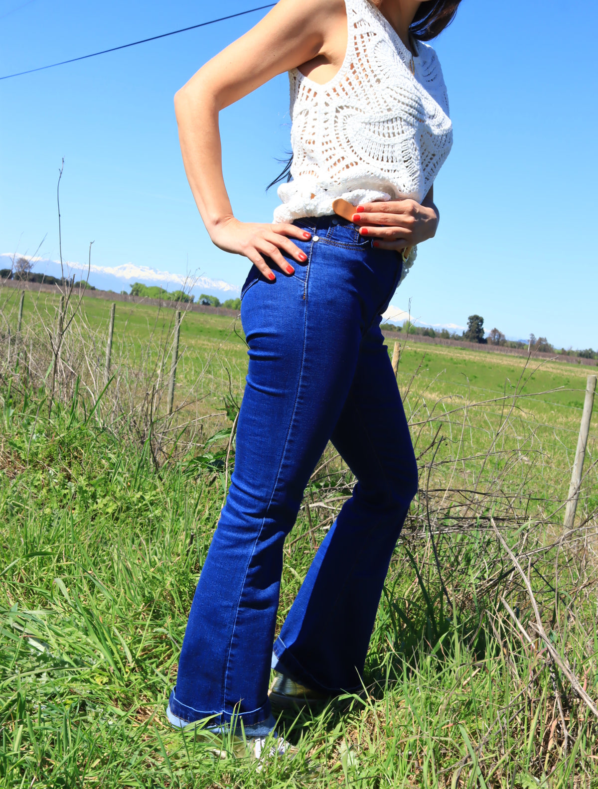 Mujer con jeans flare azul de tiro alto y top tejido blanco, de perfil en un campo