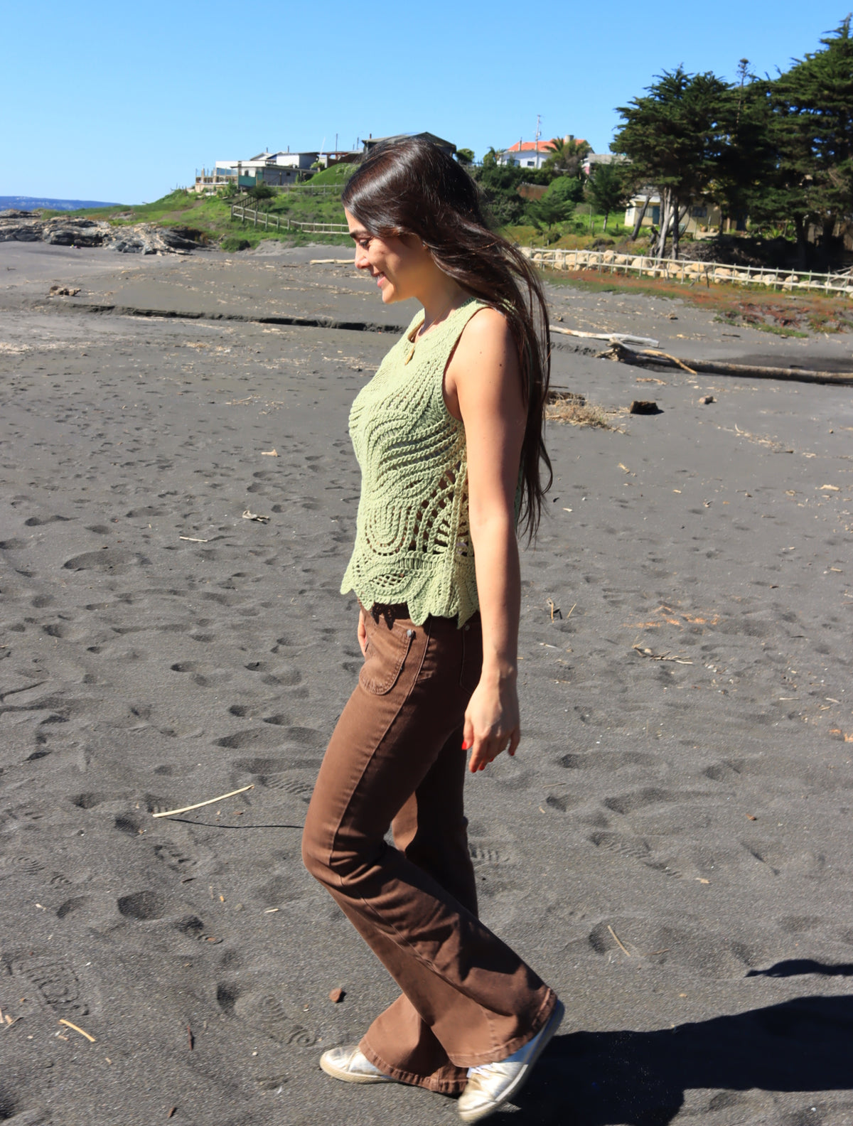 Jeans marrón de perfil, caminando en la playa con top tejido verde
