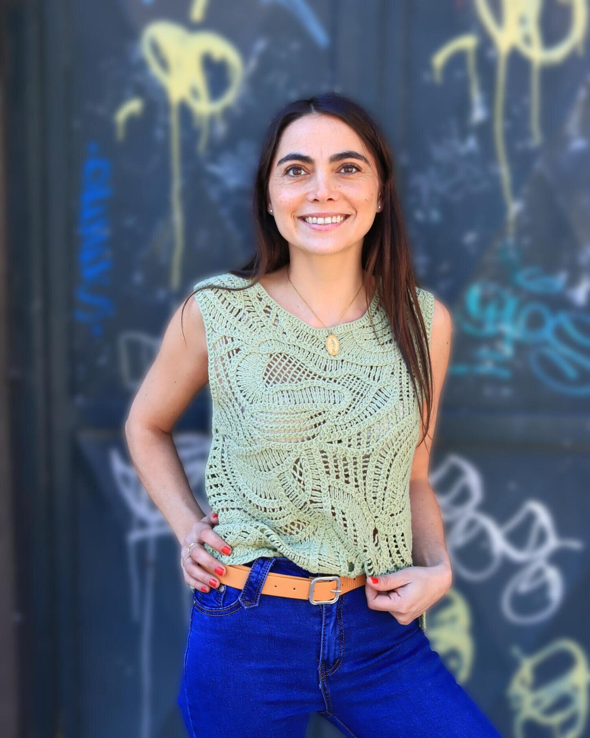 Valeria sonriendo con top crochet verde y jeans frente a un muro con grafiti de colores